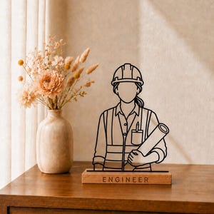 May include: A black line art silhouette of an engineer wearing a hard hat and a high-visibility vest, holding a rolled blueprint. The figure is mounted on a rectangular wooden base with the word ENGINEER engraved in capital letters.