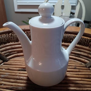 May include: A white ceramic coffee pot with a lid and a rounded handle. The pot has a long spout and a spherical knob on the lid. The pot is sitting on a woven wicker tray.