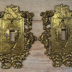 May include: A pair of gold-colored metal light switch plates featuring an ornate, embossed scrollwork border and a central rectangular panel with a shield-shaped crest at the top and bottom. Each plate has a single vertical toggle switch opening.