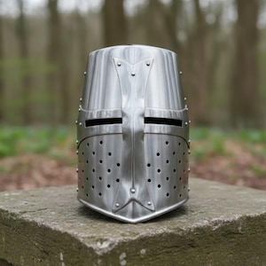 Op de afbeelding: Een gepolijste stalen kruisvaarderhelm met een platte bovenkant, geklonken constructie en een kruisontwerp. De helm heeft oogspleten en talrijke ventilatiegaten. Het wordt getoond op een stenen oppervlak.