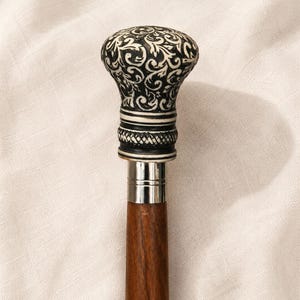 Può includere: Un bastone da passeggio con un fusto in legno marrone scuro e un collare in metallo color argento. L'impugnatura ha una forma arrotondata e bulbosa con un intricato motivo floreale a volute bianco e nero e una fascia testurizzata alla base.