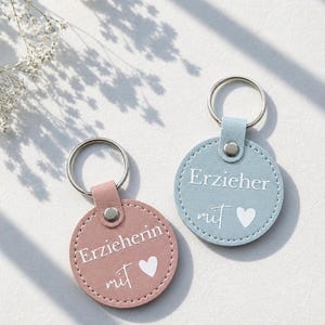 Könnte beinhalten: Zwei runde Schlüsselanhänger, einer rosa und einer blau, mit silbernen Schlüsselringen. Der rosa Schlüsselanhänger trägt die Aufschrift "Erzieherin mit" und der blaue Schlüsselanhänger "Erzieher mit", beide mit einem Herzsymbol. Die Schlüsselanhänger liegen auf einer weißen Oberfläche.