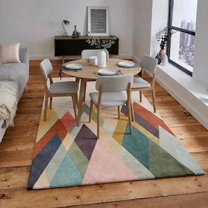 May include: A dining room scene featuring a round wooden table with four chairs, set on a colorful geometric rug. The rug has a mountain-like design in shades of pink, orange, blue, and green. A gray sofa is visible on the left.