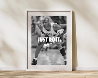 May include: Monochrome framed print of a basketball player in a dynamic pose. The image displays the text "JUST DO IT" and the Nike logo. The player is in a jersey and shorts, with a knee support. The frame is white.