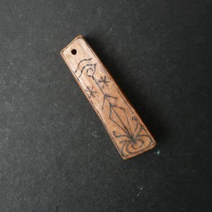 May include: A rectangular wooden pendant with a light brown finish and a small hole at the top. The surface features a dark, hand-drawn linear design consisting of a central diamond shape, swirling lines at the base, and two small star symbols.