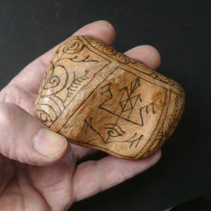 Könnte beinhalten: Ein hellbraunes, unregelmäßig geformtes Holzobjekt mit einer glatten, polierten Oberfläche. Es ist mit dunklen, handgezeichneten Tintenillustrationen versehen, darunter geometrische Muster, geschwungene Linien und ein zentrales Symbol.