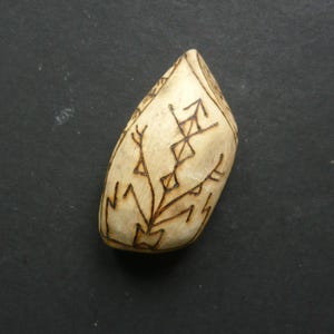 May include: A light-coloured, teardrop-shaped wooden pendant with dark brown etched designs. The pendant features geometric and branch-like patterns. The background is a dark grey surface.