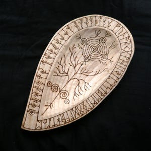 May include: A wooden, teardrop-shaped bowl with intricate pyrography designs. The light-colored wood features a central spiral, tree-like branches, and runic symbols around the edge. The bowl is likely hand-carved and suitable for decorative use.