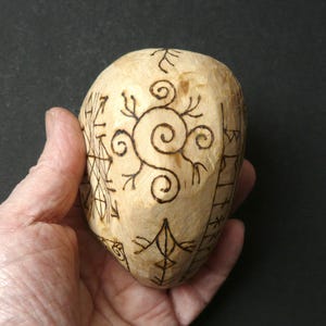 May include: A light-colored, carved wooden stone with intricate dark brown etched designs. The stone features a central spiral motif and various geometric and linear patterns, held in a hand against a dark background.