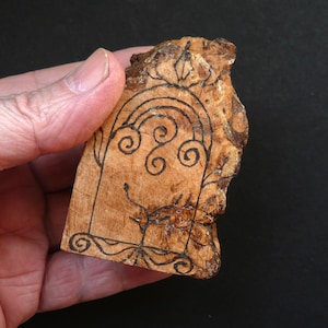 May include: A piece of light brown wood with a rough, natural edge on the right side. The surface features a hand-drawn black ink illustration of an arched doorway or window frame with decorative scrollwork and a small plant design at the base.