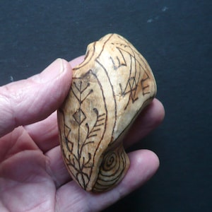 May include: A hand-carved wooden object with etched designs. The light wood has dark brown lines creating geometric and floral patterns. A spiral design is on the bottom. The object is held in a hand. The measurements are in centimeters.
