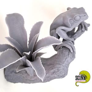 Op de afbeelding: Een monochroom grijs 3D-geprint beeldje van een kikker op een getextureerde boomtak naast een stekelige vetplant. De basis van de tak bevat een klein logo met een groene hagedisafbeelding en de tekst SCINK creation.