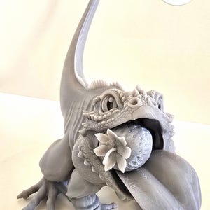 Puede incluir: Escultura gris impresa en 3D de una criatura parecida a un lagarto que sostiene una esfera y una flor en la boca. La escultura tiene texturas detalladas y una cola larga. Un logotipo con un lagarto verde está en la esquina superior derecha.