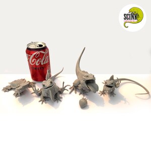 Puede incluir: Cuatro figuras de lagarto grises impresas en 3D en varias poses, incluida una con la boca abierta y una lengua larga, junto a una lata de refresco Coca-Cola Cherry roja. Un pequeño objeto gris con forma de fresa descansa cerca de las figuras.