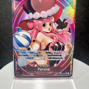 May include: A One Piece trading card featuring the character Perona with pink hair, a black top hat with white roses, and a striped plush toy. The card has a holographic finish, a cost of 4, 6000 power, and text detailing game abilities.