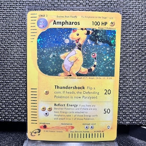 May include: A Pokémon trading card featuring Ampharos, a yellow and black creature. The card has the text "Ampharos" and "100 HP". The card is a Stage 2 Pokémon and has the text "Thundershock" and "Reflect Energy".