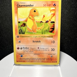 May include: A 1st Edition Charmander trading card featuring the orange lizard Pokémon in a grassy field with fire. The card displays 50 HP, the Scratch and Ember attacks, and text detailing its length of 2' 0" and weight of 19 lbs.