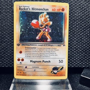 Może przedstawiać: Karta Pokémon Rocket's Hitmonchan. Karta przedstawia postać z kreskówki w pozycji bojowej, w rękawicach bokserskich i spódnicy. Karta zawiera tekst, w tym imię postaci, HP i szczegóły ataku. Karta znajduje się na przezroczystym stojaku wystawowym.