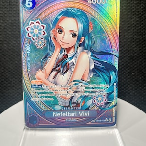Può includere: Una carta collezionabile olografica di One Piece raffigurante Nefeltari Vivi con lunghi capelli blu, che indossa un top bianco con un nastro rosso. La carta riporta il testo Nefeltari Vivi, Alabasta e meccaniche di gioco con costo 5 e 4000.