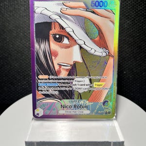 May include: A holographic One Piece trading card featuring Nico Robin from the Straw Hat Crew. The card displays a character illustration, the text 'Nico Robin', 'Straw Hat Crew', '5000', 'Banish', and 'When Attacking' with game rules in a protective case.