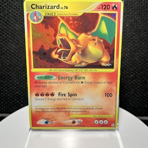 Könnte beinhalten: Eine Charizard-Sammelkarte in einer schützenden Kunststoffhülle. Die Karte hat einen gelben Rand, eine holografische Illustration eines feuerspeienden orangefarbenen Drachen und Text, darunter Charizard Lv.76, Energy Burn und Fire Spin 100.