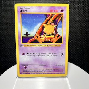 May include: A first edition Abra trading card with a yellow border and purple background. The card features a yellow creature sitting under a tree, 30 HP, and the Psyshock attack. Text includes Abra, 30 HP, Psyshock, and descriptive Pokémon data.