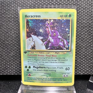 May include: A Heracross Pokémon trading card, featuring a purple beetle-like creature with large horns. The card has a yellow border and includes text detailing the Pokémon's abilities and stats. The card is displayed in a protective sleeve.