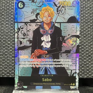 May include: A holographic trading card featuring the character Sabo from the Dressrosa/Revolutionary Army. The card has a blue and silver design with the number 6 and the text "7000". The card also includes text bubbles and a character illustration.