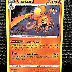 May include: A Charizard Pokémon card from the Vivid Voltage set. The card features an illustration of a Charizard with orange and yellow flames. The card has a yellow border and includes text such as "Battle Sense" and "Royal Blaze".