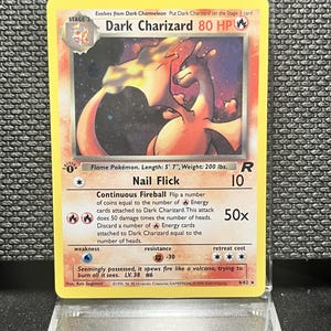 May include: A Dark Charizard trading card featuring a fire-type Pokémon illustration, 80 HP, and the Nail Flick attack. The card displays the text 'Flame Pokémon. Length: 5' 7", Weight: 200 lbs.' and includes a first edition stamp on the left side.