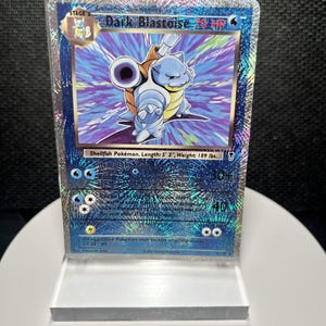May include: A Dark Blastoise trading card in a clear protective sleeve, featuring a blue character with cannons on its back, a holographic starburst background, and text including 70 HP, Shellfish Pokémon, Length 5' 3", and Weight 189 lbs.