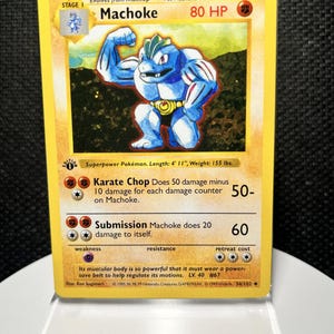May include: A yellow Machoke trading card featuring a blue muscular character with a gold belt. The card displays 80 HP, the attacks Karate Chop and Submission, and text including Superpower Pokémon, Length 4' 11", and Weight 155 lbs.
