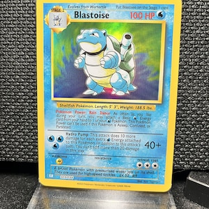 Peut inclure: Carte Pokémon Blastoise holographique. Elle présente une illustration de Blastoise bleu avec une carapace blanche et deux canons à eau. Le texte indique "Blastoise" et "100 HP". La carte est dans une protection.