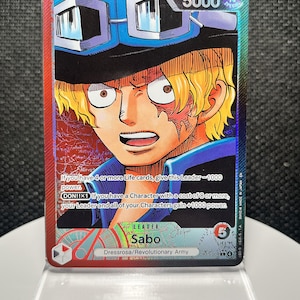 May include: A One Piece trading card featuring the character Sabo with blonde hair and goggles. The card displays a power level of 5000, text detailing game abilities, the name Sabo, and the affiliation Dressrosa/Revolutionary Army in a red and blue frame.