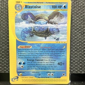 May include: A Blastoise trading card with a yellow border and blue background. The card features the Stage 2 Pokémon Blastoise, 100 HP, and the Energy Cannon attack. Text includes Poke-POWER Jet Stream, 2002 Pokemon/Nintendo, and 37/165.