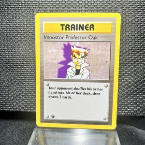 May include: A Trainer card featuring "Impostor Professor Oak" with a yellow border. The card text reads, "Your opponent shuffles his or her hand into his or her deck, then draws 7 cards." The card is displayed on a clear acrylic stand.