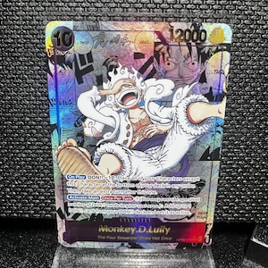 May include: A holographic trading card featuring Monkey D. Luffy from the One Piece anime. The card has a value of 12000 and features Luffy in a dynamic pose with the text "GEAR FOUR". The card is displayed on a clear acrylic stand.
