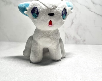 May include: A small plush toy shaped like a fox with white fur, light blue inner ears, and large embroidered eyes featuring purple and teal accents. The creature has a small black nose and a tiny red mouth, with a tuft of white fur on its head.