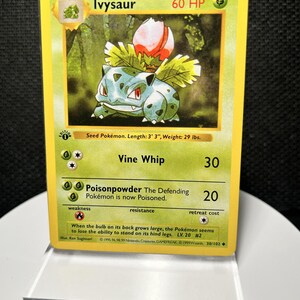 Könnte beinhalten: Eine gelbe Ivysaur-Sammelkarte mit einem grünen Wesen und einer rosa Blütenknospe auf dem Rücken. Die Karte zeigt den Text Vine Whip und Poisonpowder, mit 60 KP, einem 1st Edition-Stempel und dem Copyright-Datum 1999 am unteren Rand.