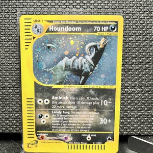 Könnte beinhalten: Eine Pokémon-Sammelkarte mit Houndoom, einem Pokémon vom Typ Finsternis. Die Karte hat einen gelben Rand und zeigt den Namen, die KP und die Angriffe des Pokémon. Die Illustration zeigt Houndoom vor einem Sternenhintergrund. Die Karte ist 12 cm lang und wiegt 35 kg.