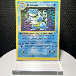 Könnte beinhalten: Eine holografische Blastoise-Sammelkarte mit einer blauen, schildkrötenartigen Kreatur mit Kanonen auf dem Rücken. Die Karte zeigt 100 KP, die Kraft Regentanz und die Attacke Hydropumpe. Es handelt sich um eine 1st Edition Karte der Stufe 2.