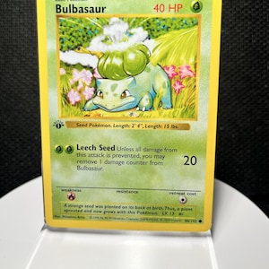 May include: A 1999 Bulbasaur trading card with 40 HP, featuring a green creature with a bulb on its back. The card text includes 'Seed Pokémon. Length: 2' 4", Length: 15 lbs.' and the Leech Seed attack description. It is encased in a clear plastic sleeve.