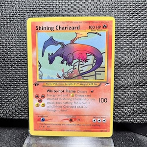 Op de afbeelding: Een Shining Charizard Pokémon-kaart met een paarse en rode drakenillustratie. De kaart heeft een gele rand en bevat tekst zoals "100 HP" en "White-hot Flame". De kaart is 170 cm lang en weegt 91 kg.