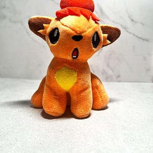 May include: A plush toy of a small orange fox-like creature with large dark eyes, a yellow chest patch, and a bright red tuft of hair on its head. The creature has pointed brown-tipped ears and an open mouth expression, crafted from soft fabric.