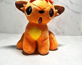 May include: A plush toy of a small orange fox-like creature with large dark eyes, a yellow chest patch, and a bright red tuft of hair on its head. The creature has pointed brown-tipped ears and an open mouth expression, crafted from soft fabric.