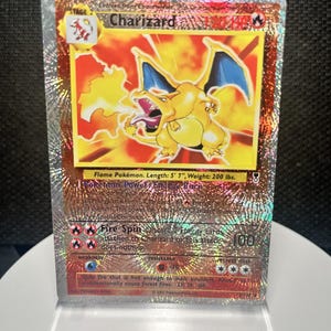 May include: A holographic Charizard trading card featuring a yellow dragon-like creature breathing fire. The card displays 120 HP, the Flame Pokémon description, Energy Burn power, and Fire Spin attack text, all set against a shimmering, reflective background.