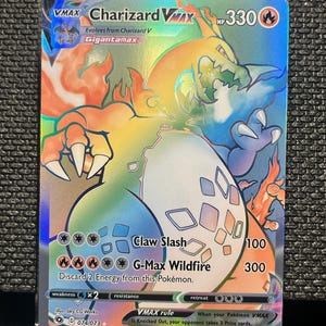 May include: A holographic Charizard VMAX Pokémon card. The card features a fiery orange and blue dragon-like creature with the text "Charizard VMAX" and "Gigantamax". The card has a HP of 330 and includes attack details.