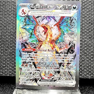May include: A holographic Charizard ex Pokémon card. The card features a fiery orange dragon with blue and green accents. Text includes "Charizard ex", "Infernal Reign", and "Burning Darkness". The card is displayed in a clear protective case.