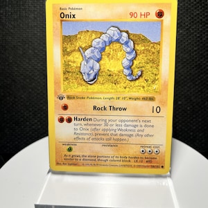 May include: A 1999 Wizards of the Coast Onix trading card featuring a yellow border, 90 HP, and the Rock Throw and Harden attacks. The card displays the Rock Snake Pokémon illustration, a first edition stamp, and text detailing its stats.
