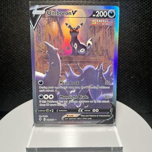 May include: A holographic Umbreon V trading card featuring a dark purple and black creature with glowing red eyes standing in a city street at night. The card displays the text Umbreon V, 200 HP, Single Strike, Mean Look, and Moonlight Blade.